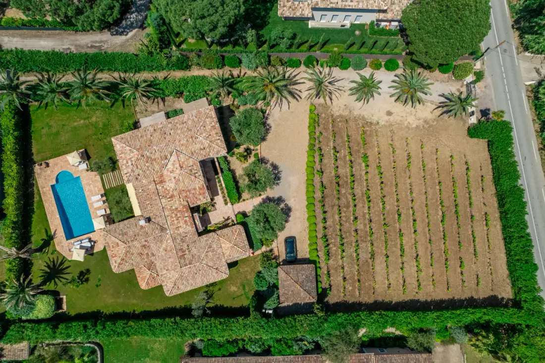 Vista aérea de una casa, una piscina y hileras cultivadas junto a una carretera.