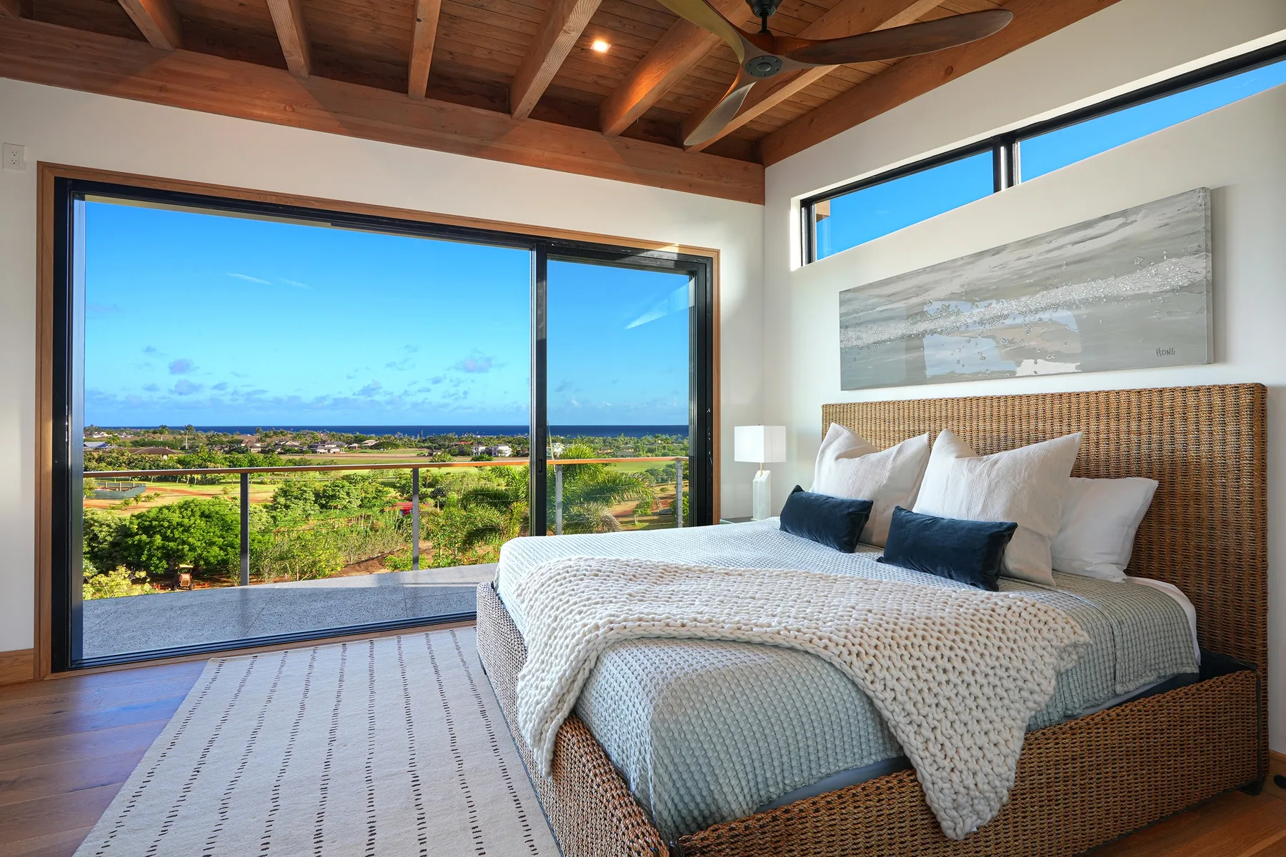 A bedroom with a large bed beside wide windows overlooking greenery and the ocean.
