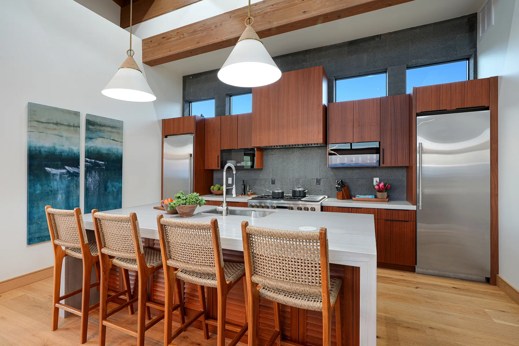 A modern kitchen with a long island and woven-back stools beneath pendant lights.
