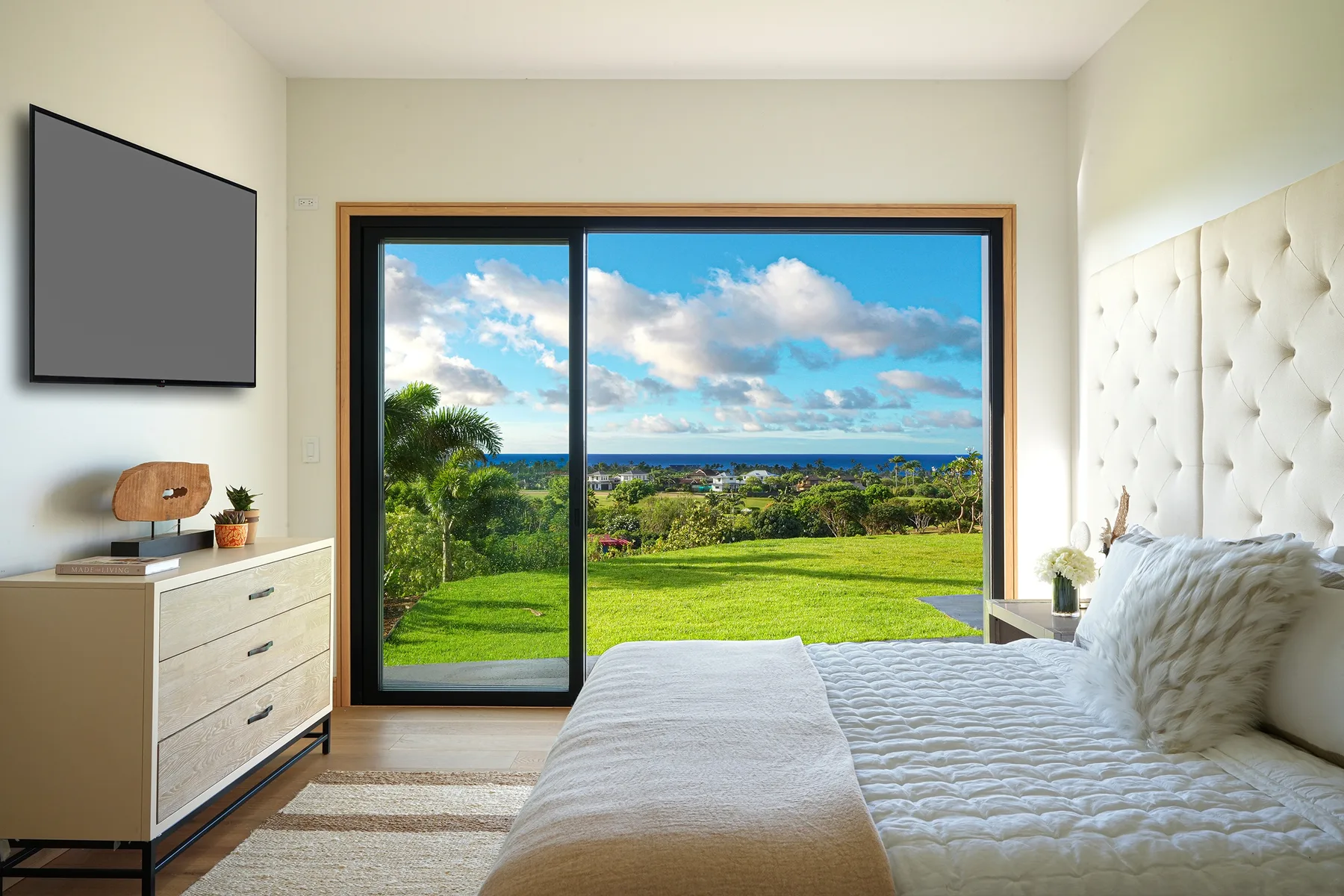 A bright bedroom with a bed beside a large window overlooking green grounds and the ocean.