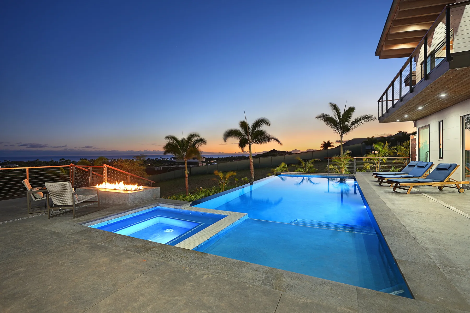 An outdoor swimming pool beside a house, with palm trees and a lit fire feature.