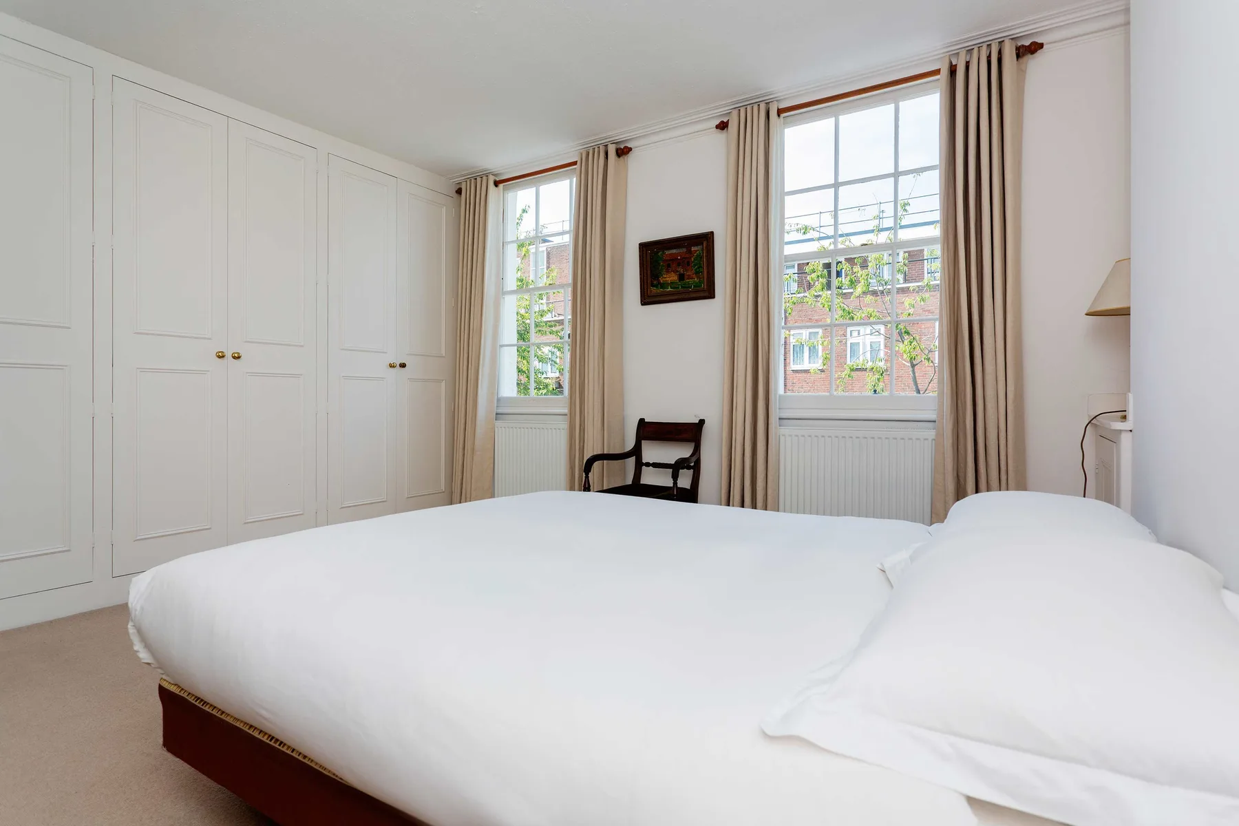 A bright bedroom with a large bed, built-in cupboards, and tall curtained windows.
