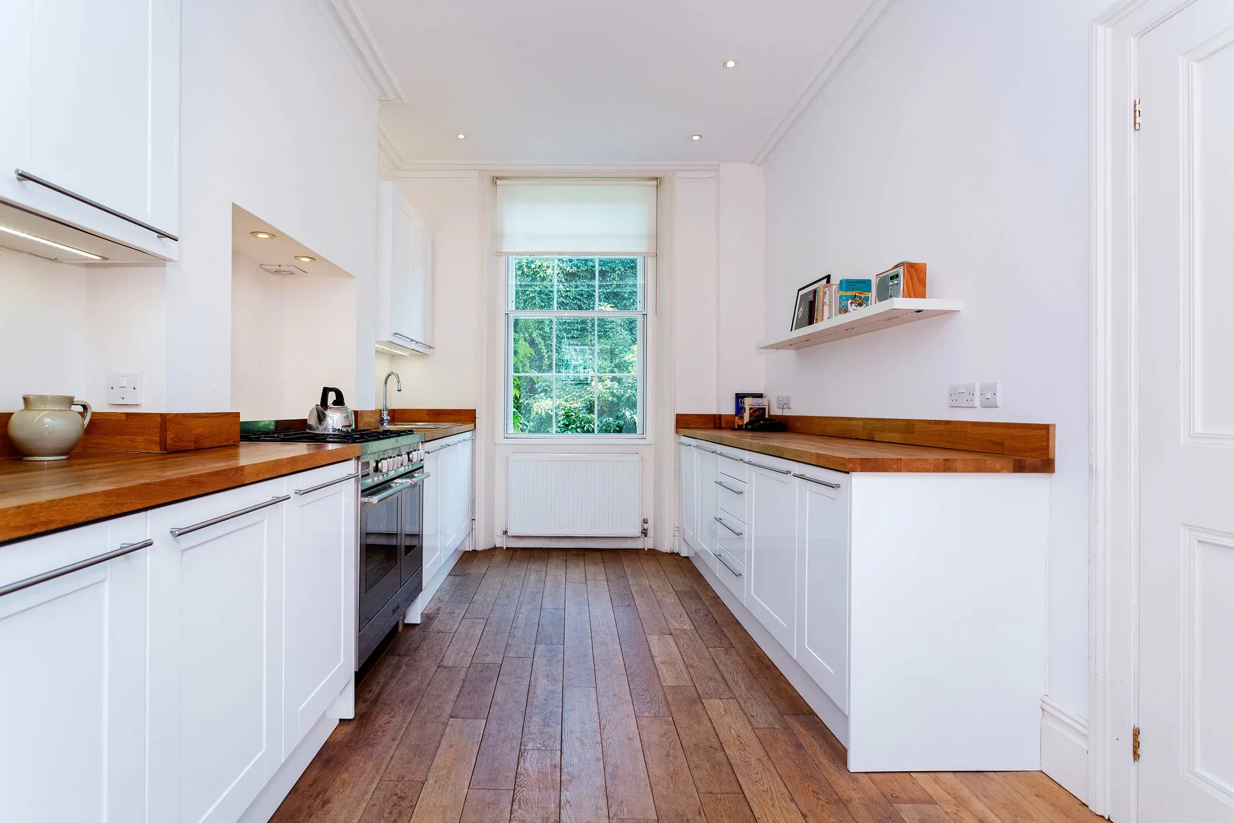 A bright galley kitchen with white cabinets, wooden counters, and a window.