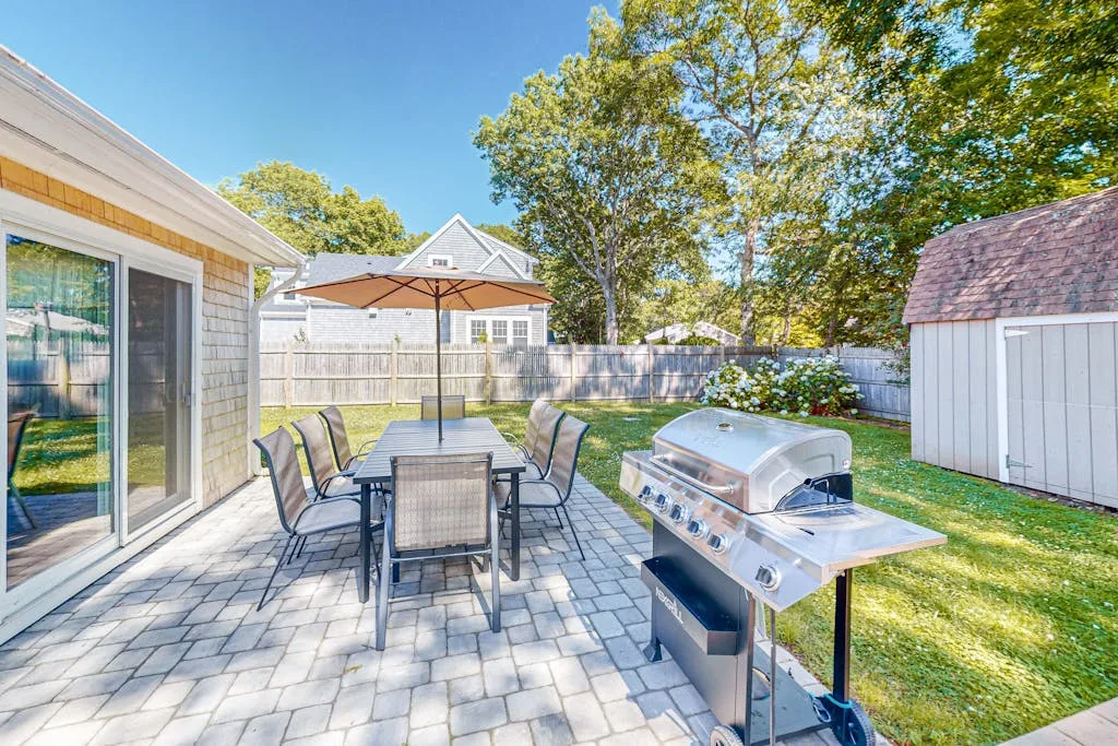 Un patio trasero pavimentado tiene asientos al aire libre, una mesa y una parrilla.