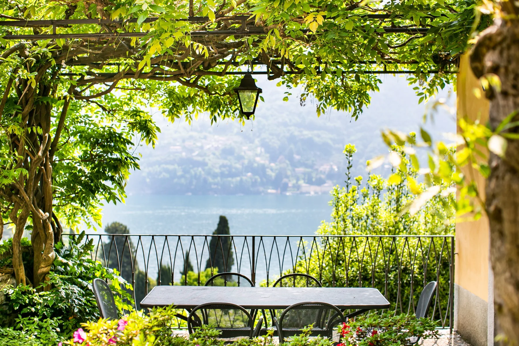Una terraza ajardinada con una mesa de comedor tiene vistas a un lago entre enredaderas frondosas.