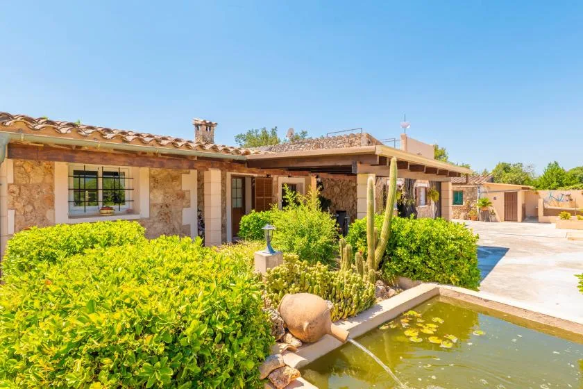 A stone house opens onto a sunny courtyard with greenery and a small pond.