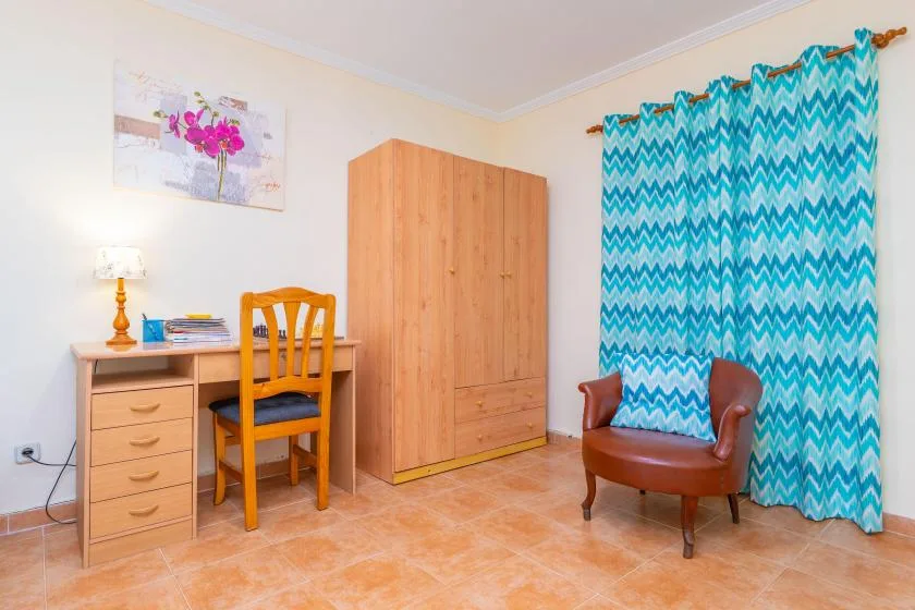 A bedroom with a desk, wooden wardrobe, and armchair beside patterned curtains.
