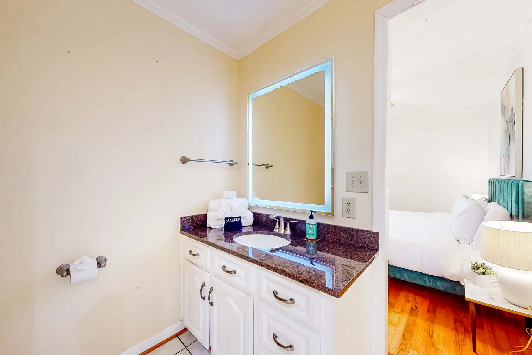 A bathroom vanity with a lit mirror and a doorway opening onto a bedroom.
