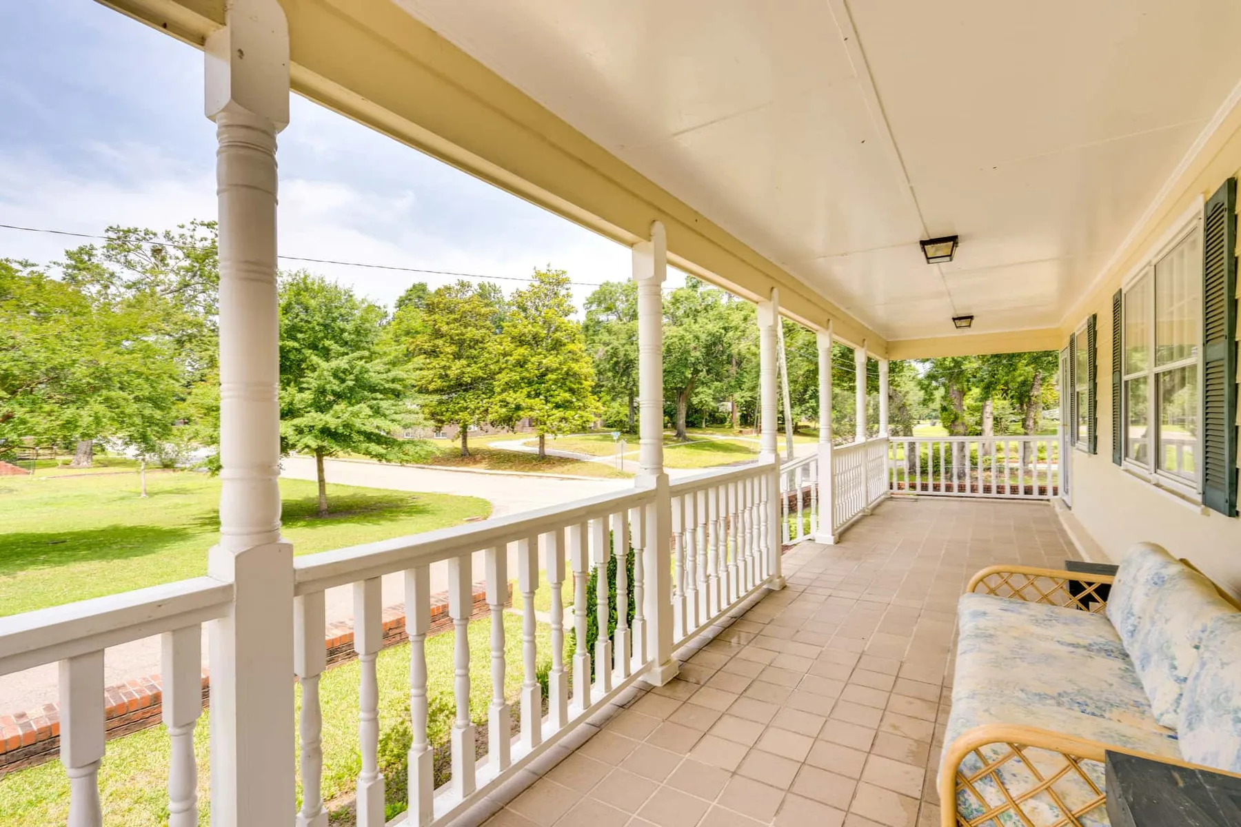 A covered porch with a railing and cushioned seating overlooks a green lawn.