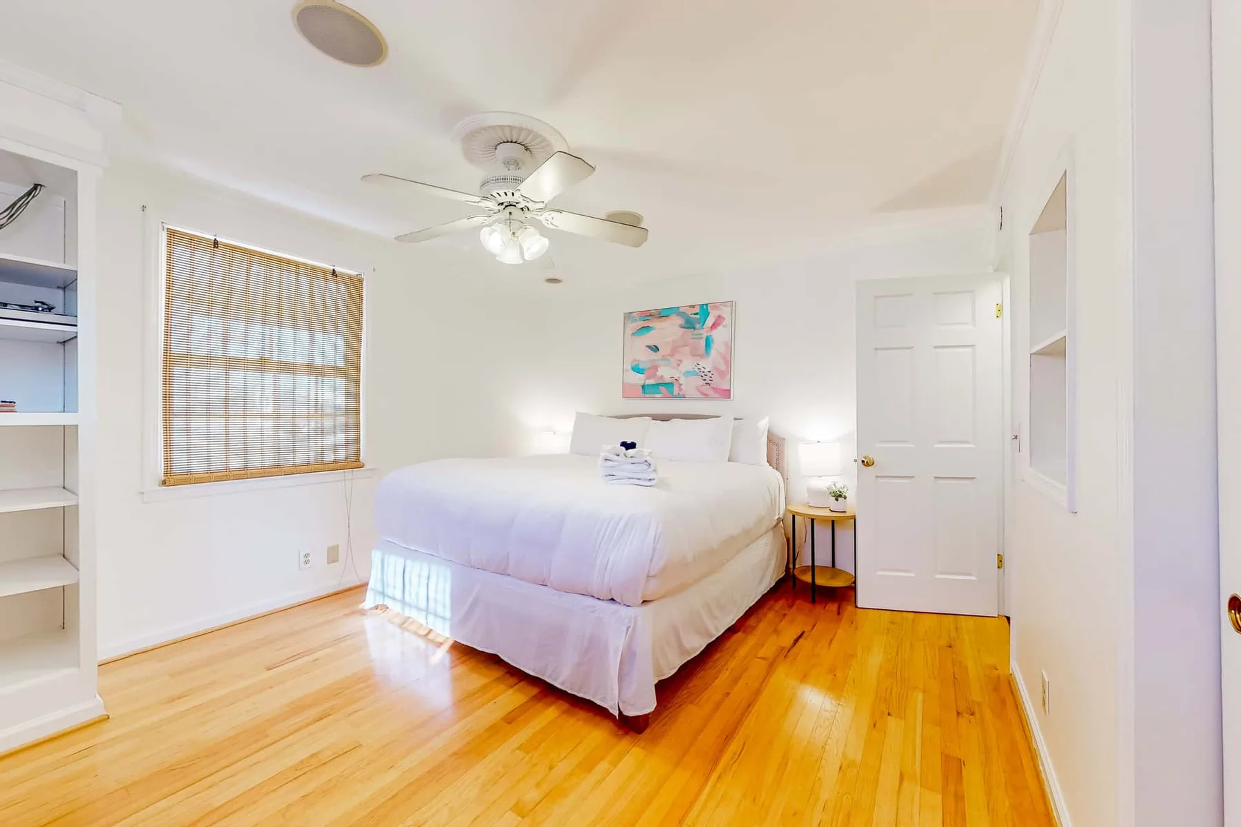A bright bedroom with a neatly made bed, ceiling fan, and wooden flooring.