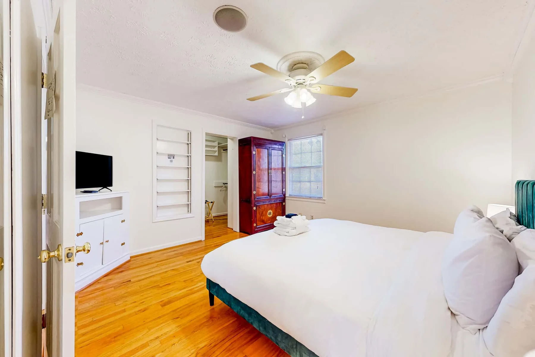 A bright bedroom with a large bed, wooden flooring, and a ceiling fan.