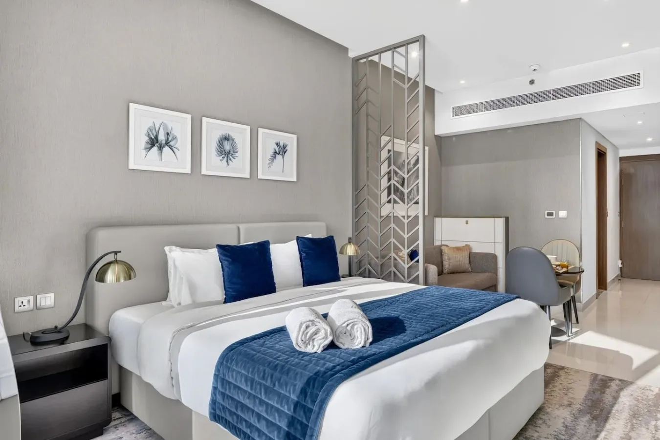 Dormitorio con una cama bien tendida, detalles azules y láminas botánicas enmarcadas.
