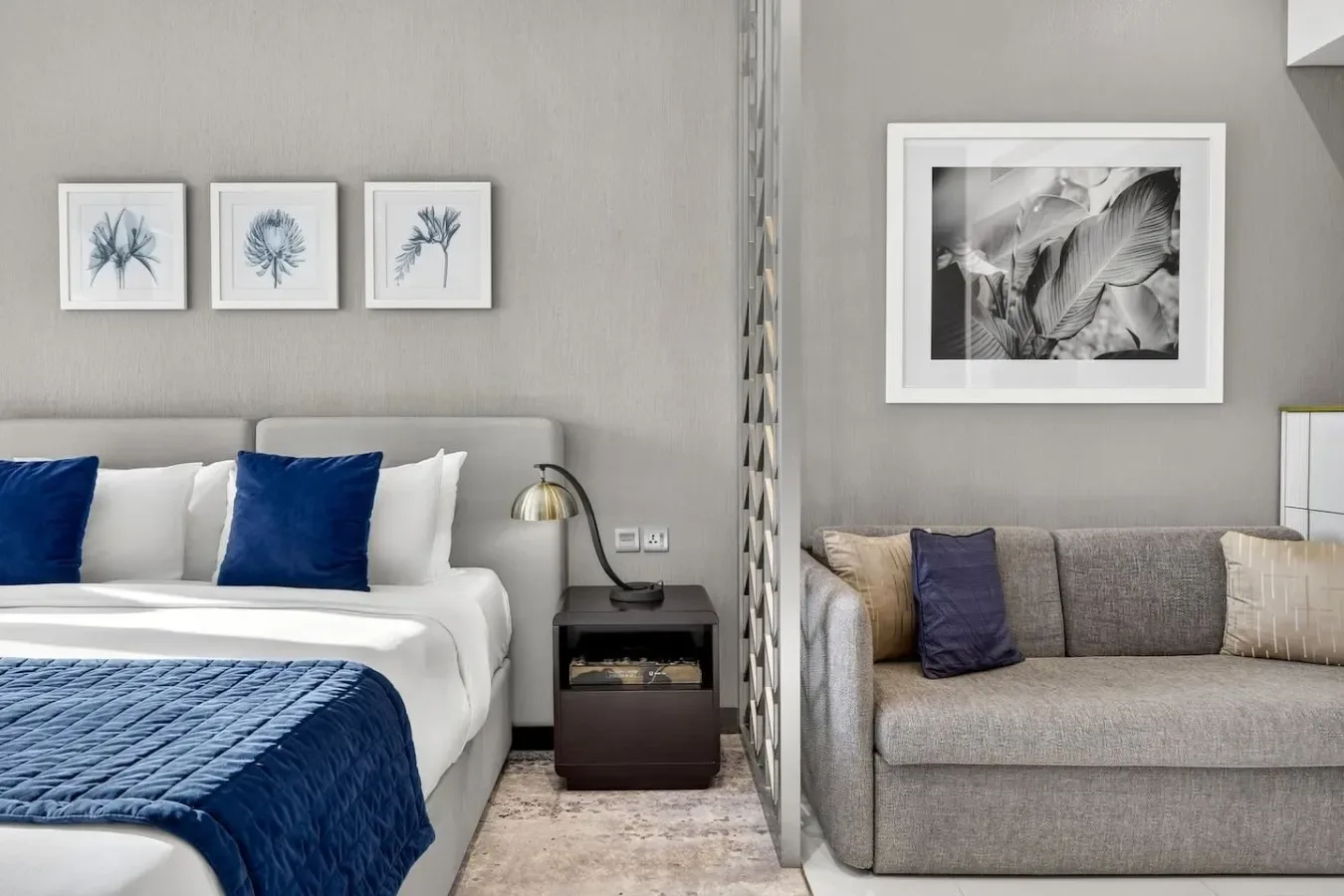 Dormitorio y zona de estar con una cama, un sofá gris y láminas botánicas enmarcadas.
