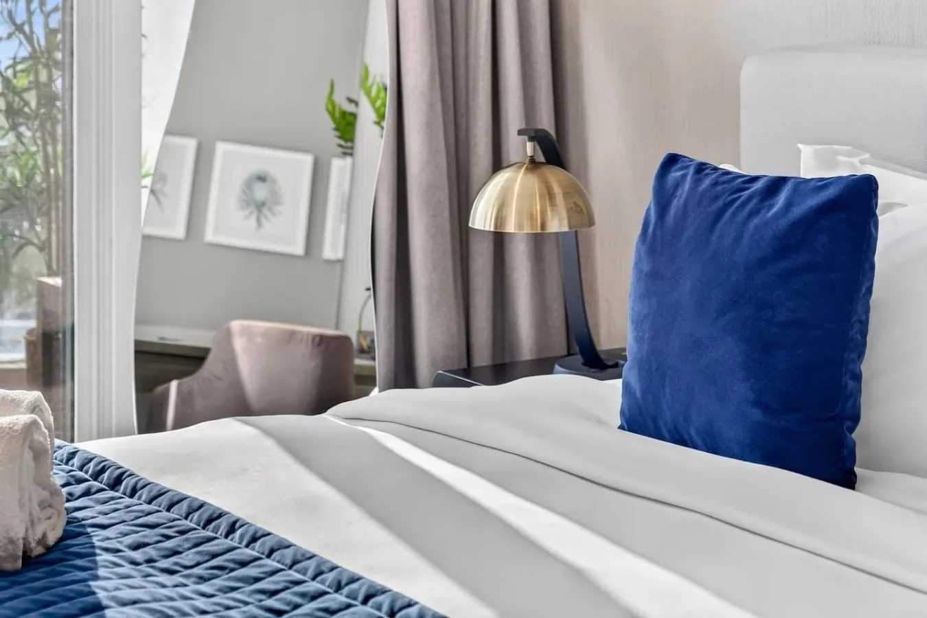 Una cama bien tendida con ropa de cama azul está junto a una lámpara y cortinas grises.