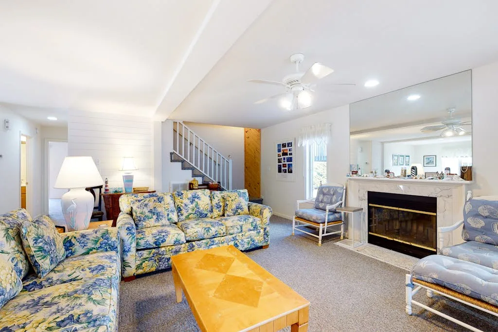 A bright living room with floral seating, a fireplace, and a wooden coffee table.