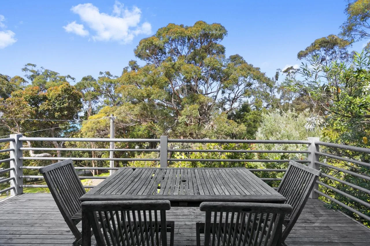 An outdoor deck has a dark slatted dining table and seating surrounded by trees.