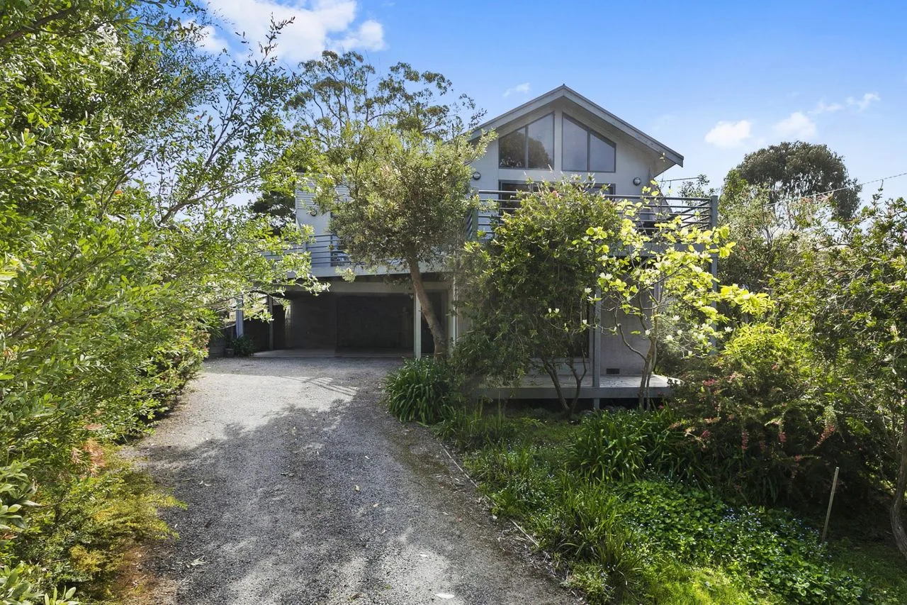 A two-story house stands at the end of a gravel driveway, surrounded by greenery.