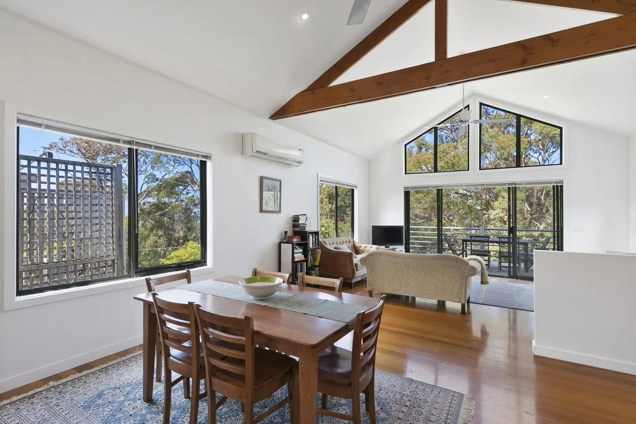 A bright open-plan living and dining area with large windows and exposed ceiling beams.