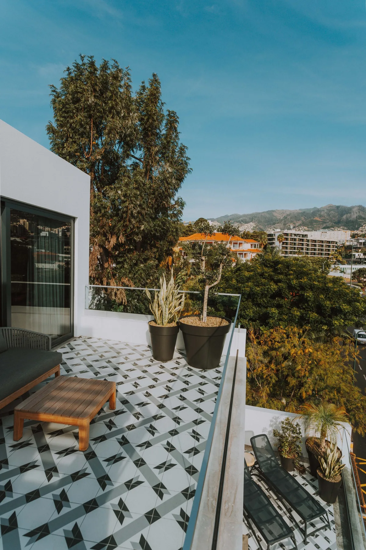 Una terraza exterior con suelo de baldosas, asientos y plantas en macetas da a una ladera verde.