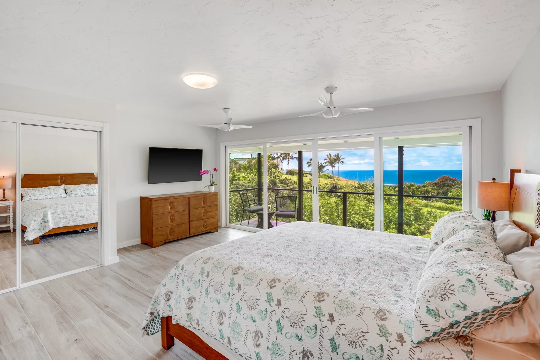 Un dormitorio con cama, cómoda y amplias ventanas con vistas a vegetación y al océano.