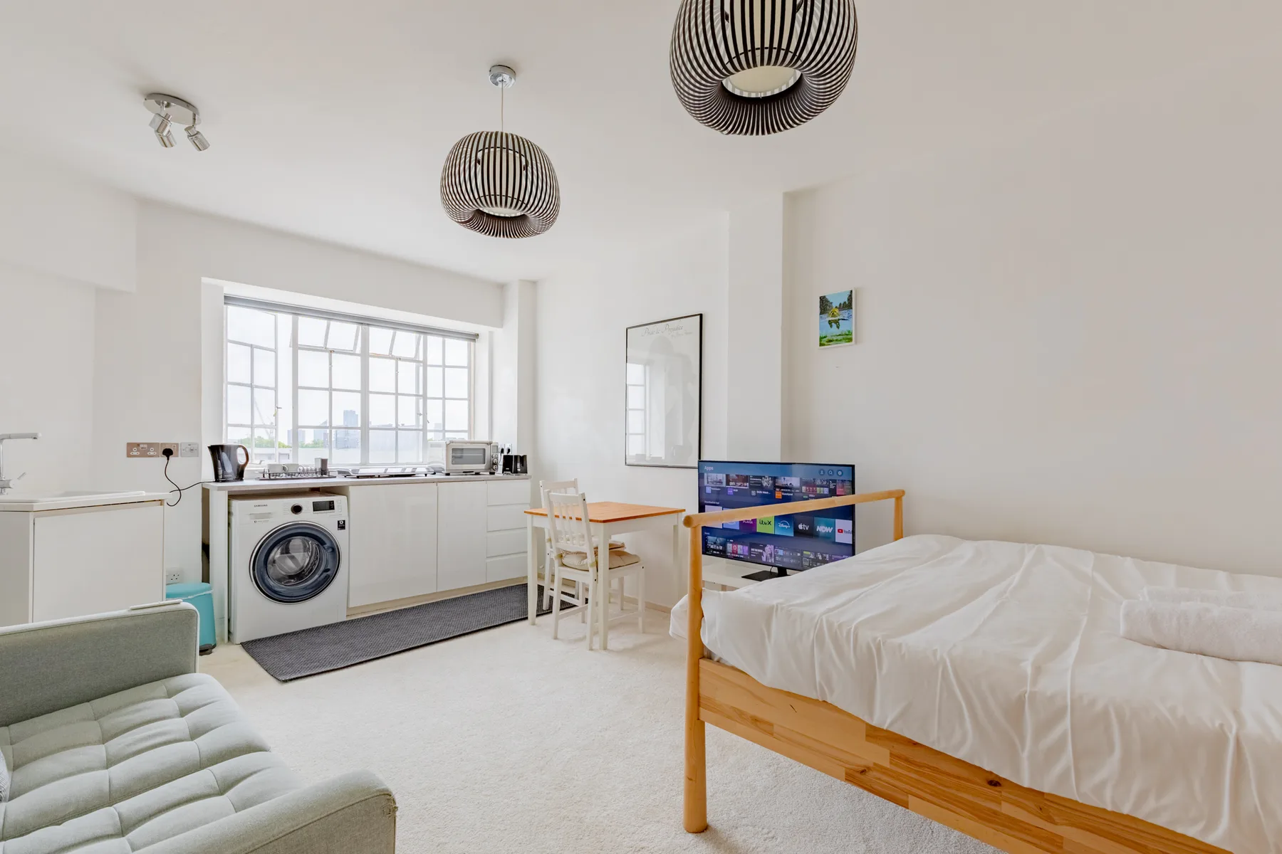 A bright room with a bed, seating, and a kitchenette by the windows.