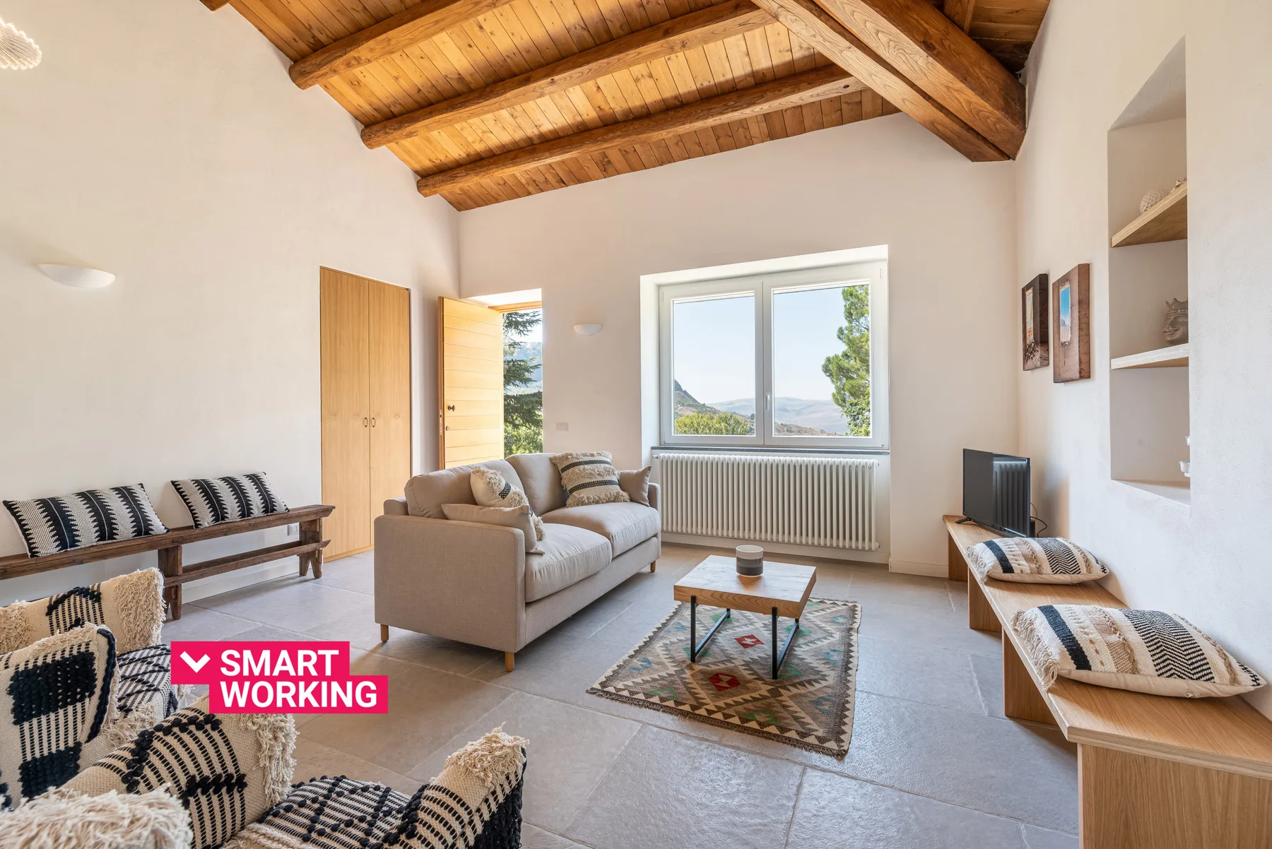 A bright living room with seating, a coffee table, and a large window beneath a wooden ceiling.