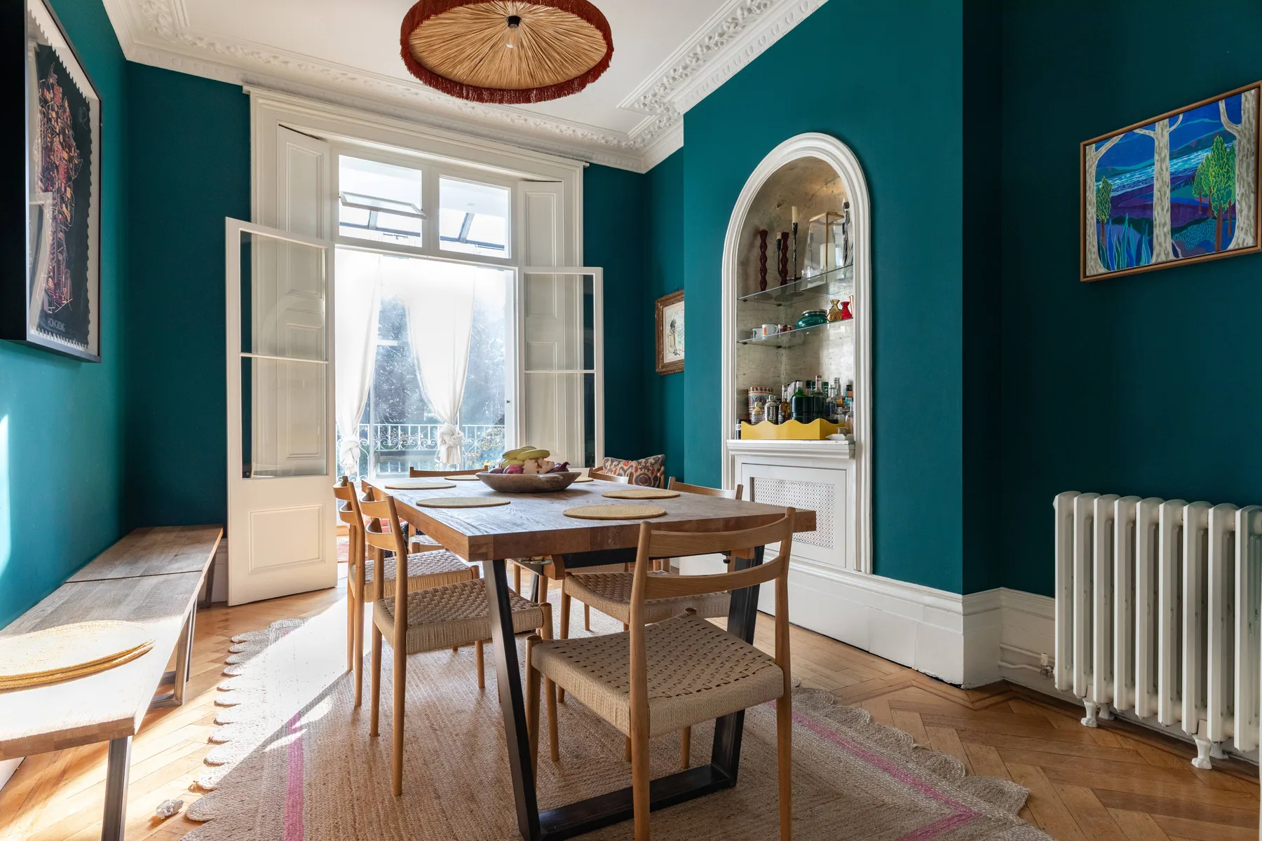A dining room with a wooden table and chairs beside tall windows.