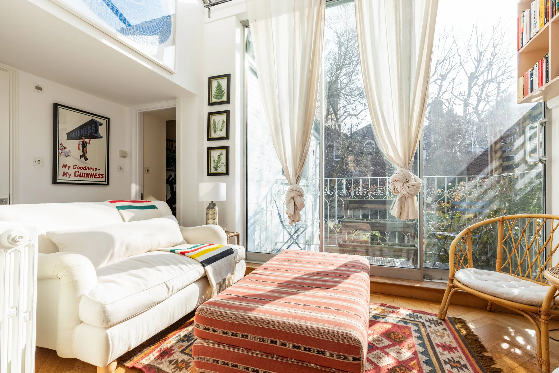 A bright living room with seating, a patterned ottoman, and tall windows opening onto a balcony.
