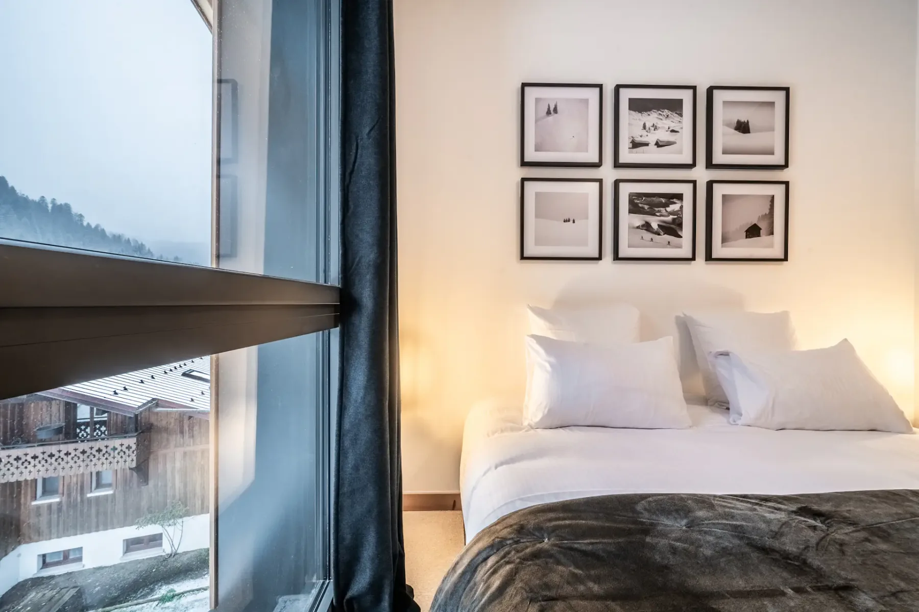 Dormitorio acogedor con una cama doble, almohadas blancas y una manta oscura. A la izquierda hay una ventana con cortinas, y sobre la cama hay cuadros enmarcados dispuestos en cuadrícula en la pared.