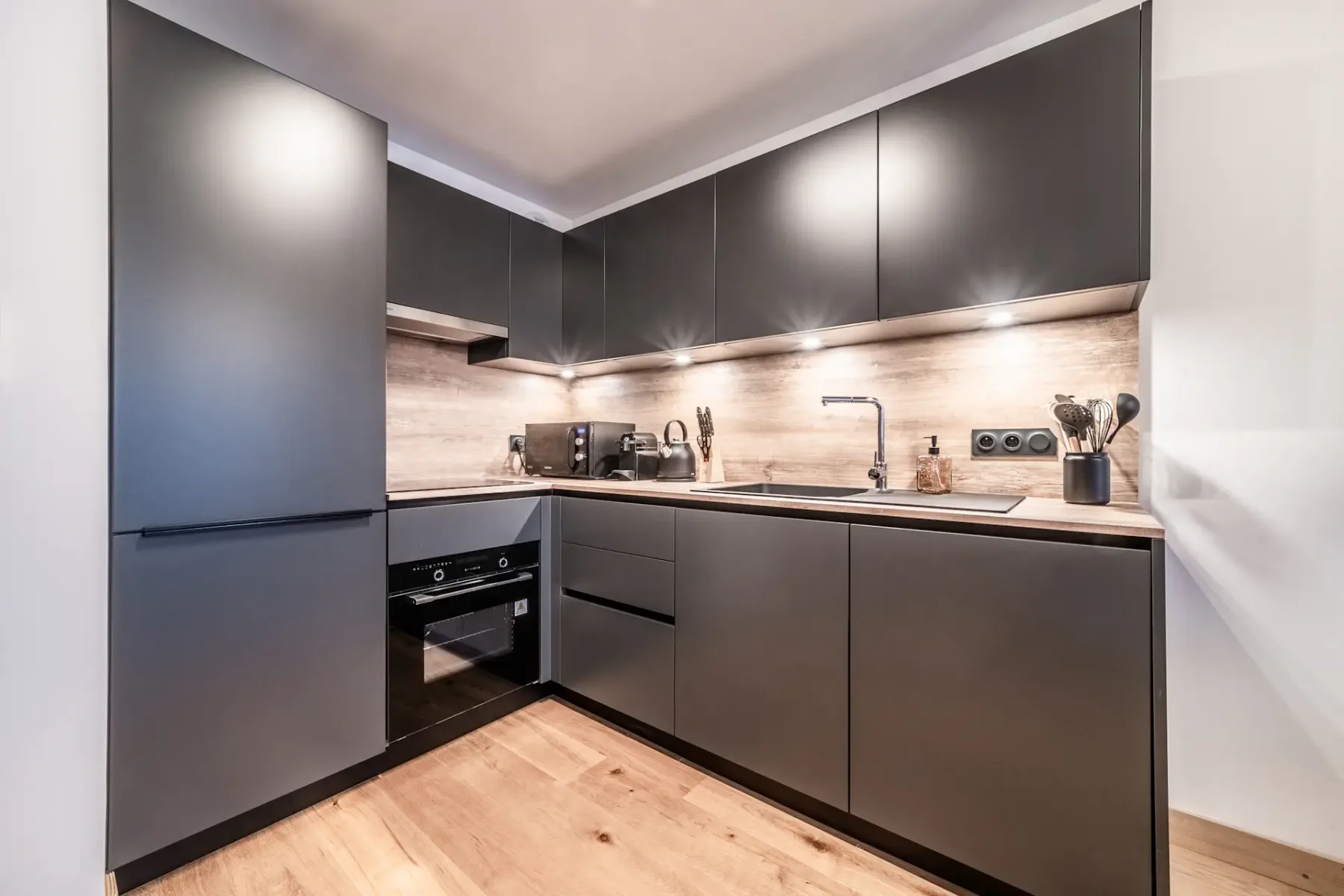 Cocina moderna con armarios oscuros, encimera de madera y electrodomésticos integrados. Una intensa iluminación bajo los armarios ilumina el espacio, que cuenta con fregadero, cocina y una decoración minimalista.