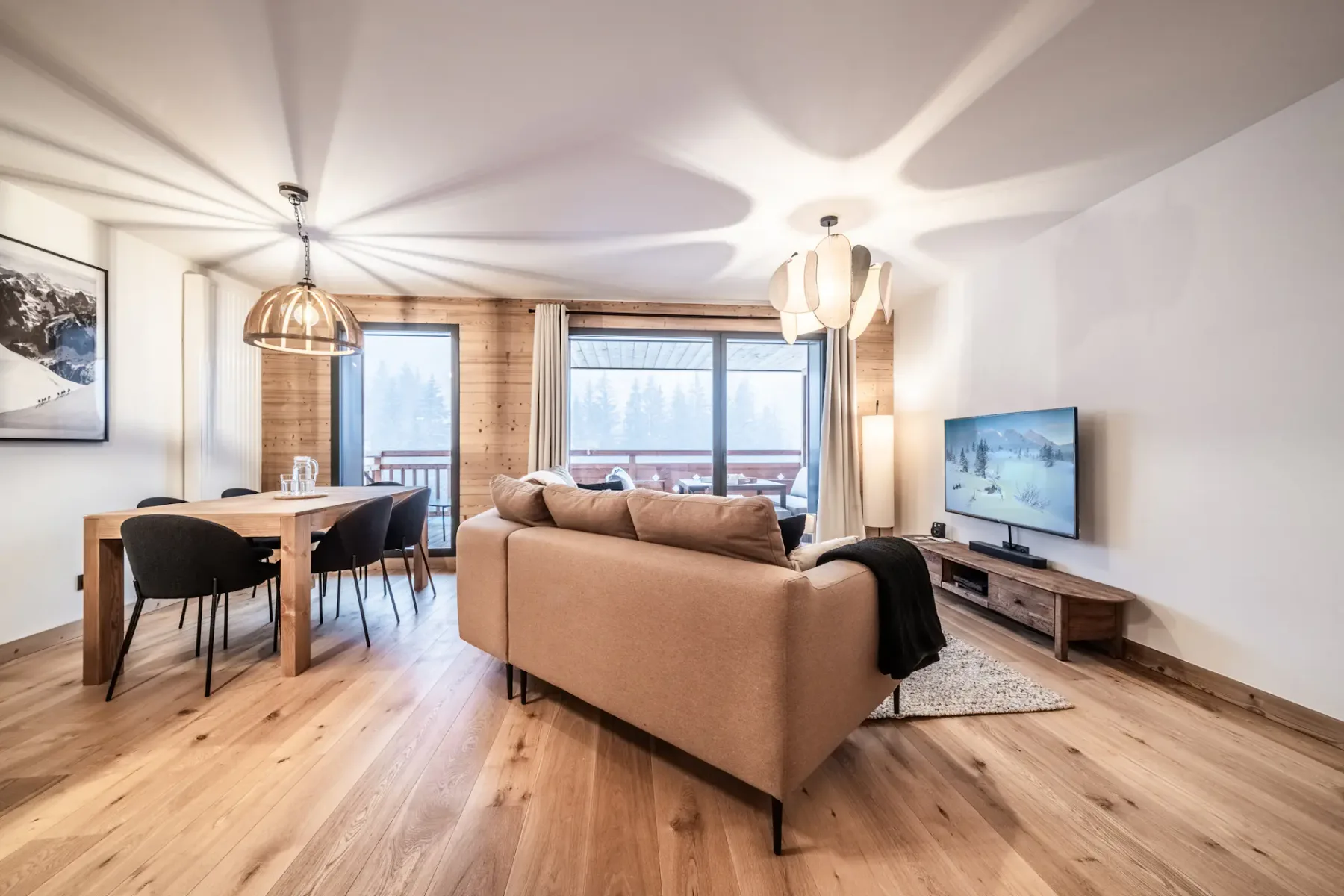 Sala de estar moderna con un sofá de color marrón claro, una mesa de comedor de madera, sillas negras y un televisor. Las grandes ventanas dejan entrar luz natural, que resalta los suelos y las paredes de madera.