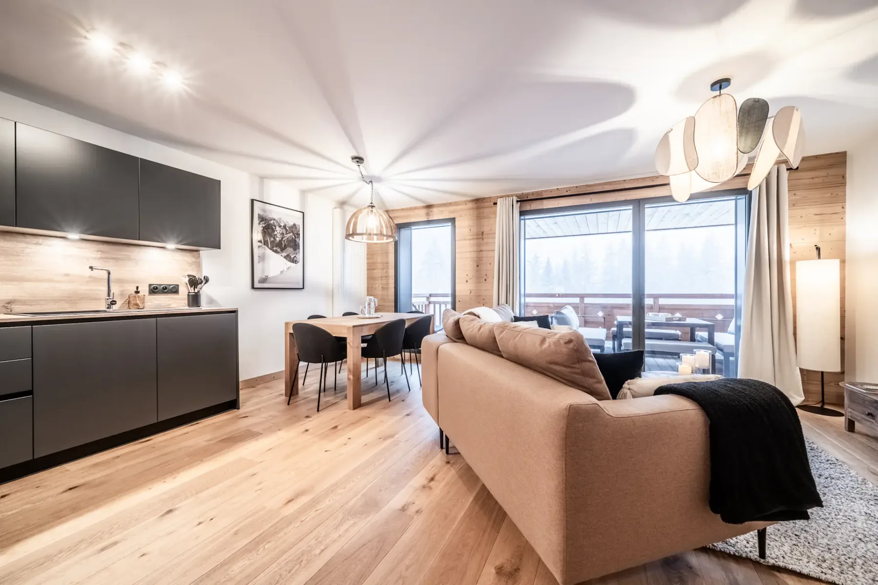 Sala de estar y cocina modernas de planta abierta con suelos de madera, armarios negros, sofá marrón, mesa de comedor y grandes ventanales que dan a un balcón con vistas a los árboles.