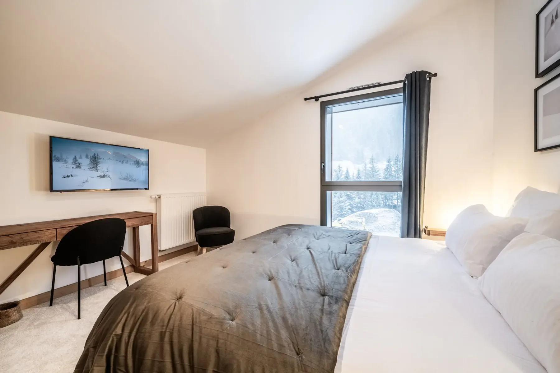Dormitorio acogedor con una cama, un escritorio, una silla y un televisor montado en la pared. Una ventana grande muestra un paisaje nevado.