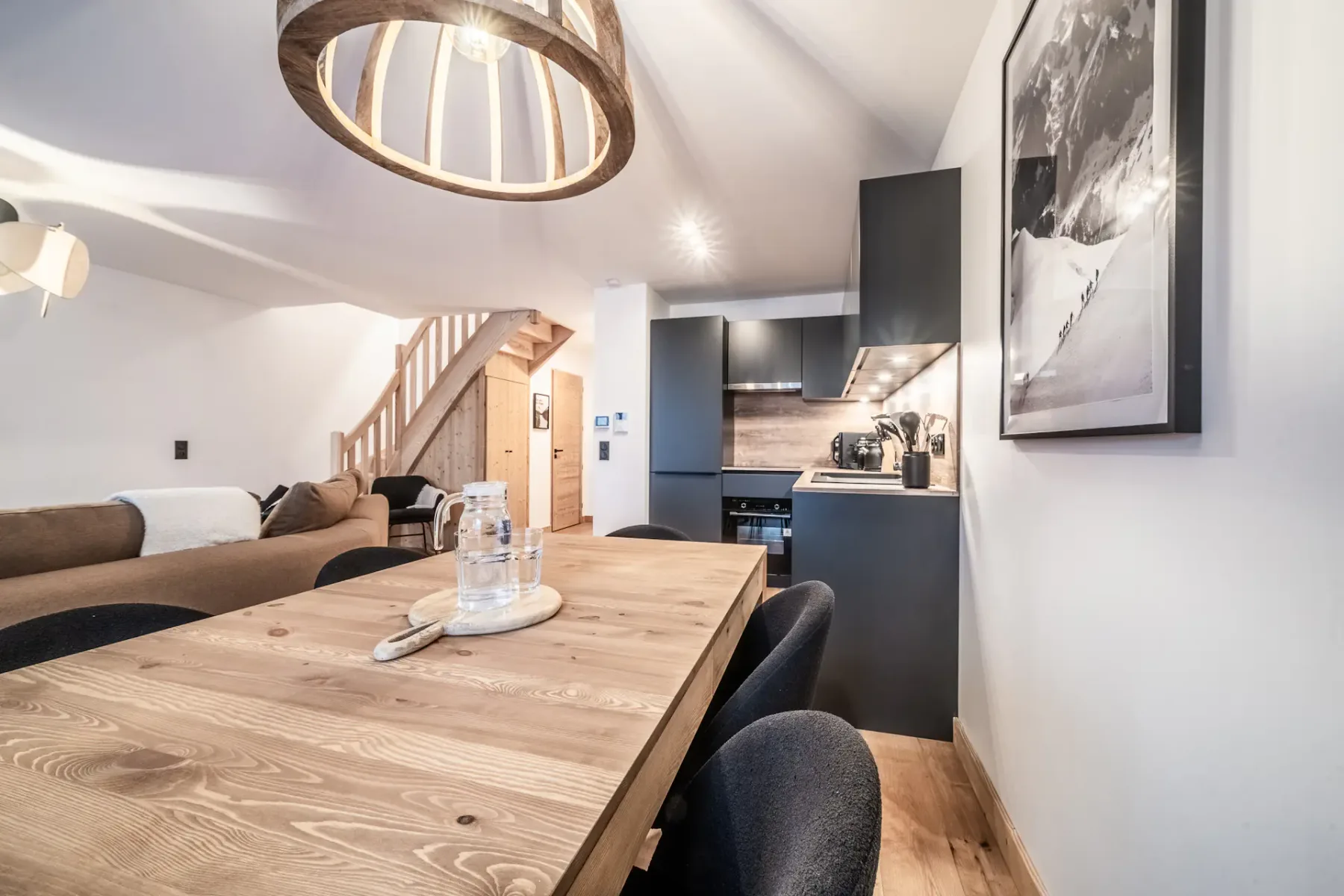 Cocina y comedor modernos con mesa de madera, sillas negras y una lámpara contemporánea. Una escalera conduce a una planta superior y una gran fotografía enmarcada decora la pared.