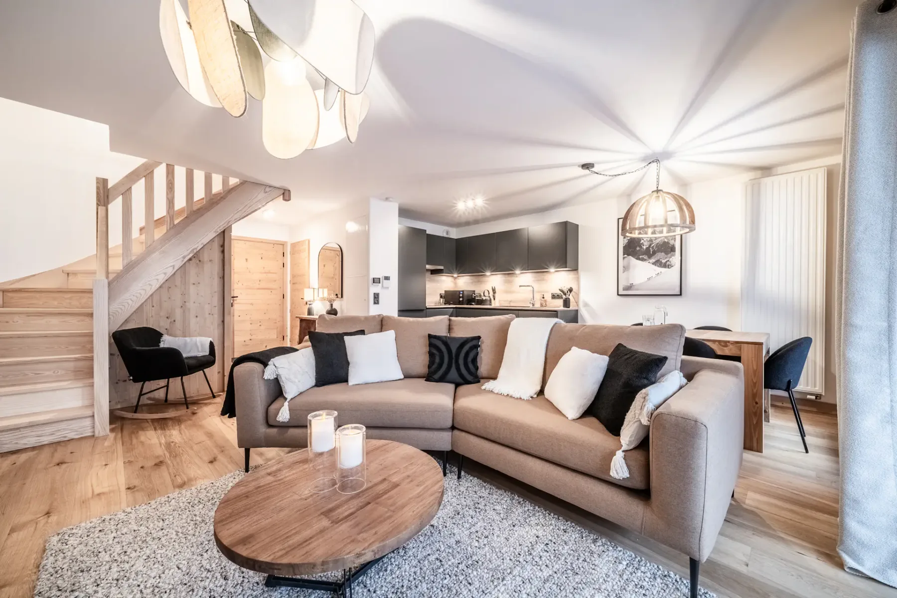 Sala de estar moderna con un sofá modular marrón, una mesa de centro de madera, velas y vistas a una cocina. La iluminación cálida y los detalles de madera crean un ambiente acogedor. Se ve una escalera a la izquierda.