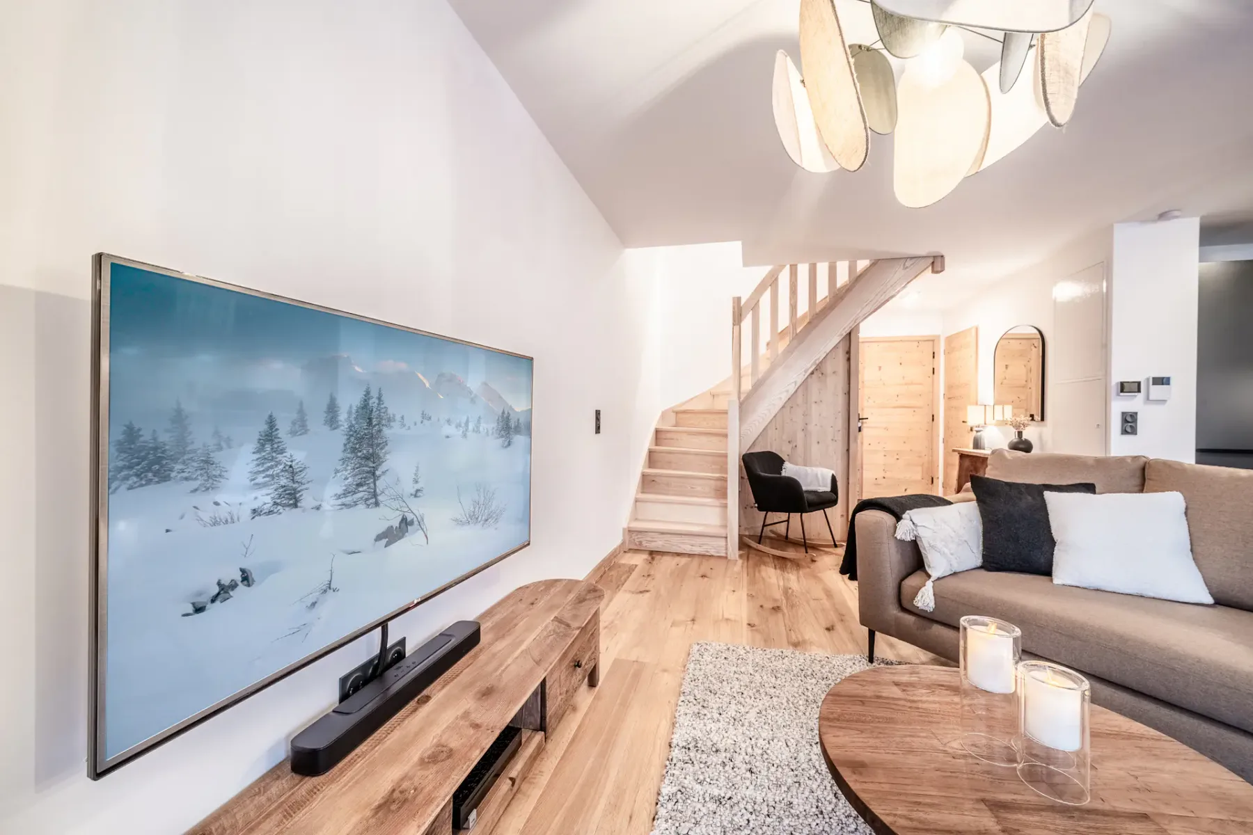 Sala de estar moderna con un televisor montado en la pared que muestra una escena nevada. Cuenta con un sofá marrón, una mesa de centro de madera, una escalera y una singular lámpara de techo. Ambiente acogedor con iluminación cálida.