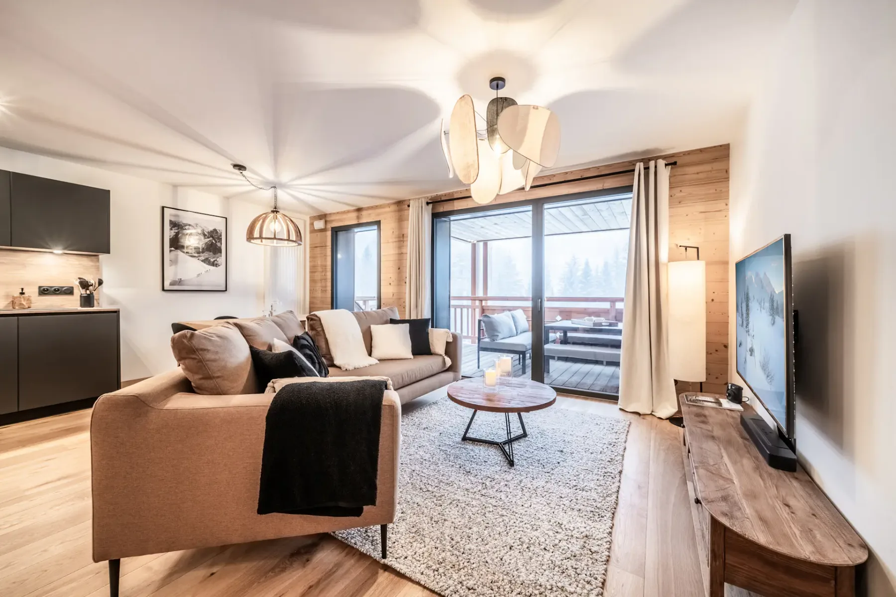 Sala de estar moderna con un sofá beige, detalles de madera, un televisor grande y puertas de vidrio que dan a una terraza de madera con asientos al aire libre y vistas a un bosque nevado.