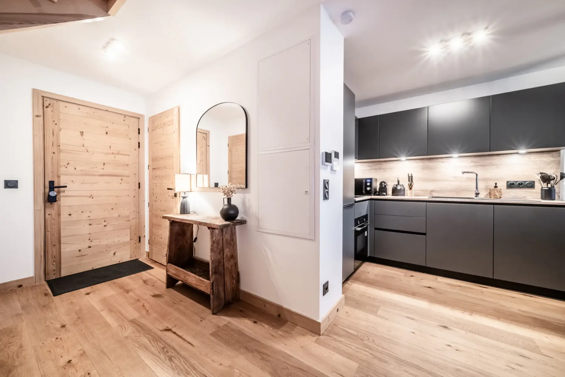 Cocina y recibidor modernos con suelos de madera, armarios oscuros, una consola de madera con un espejo redondo e iluminación ambiental.