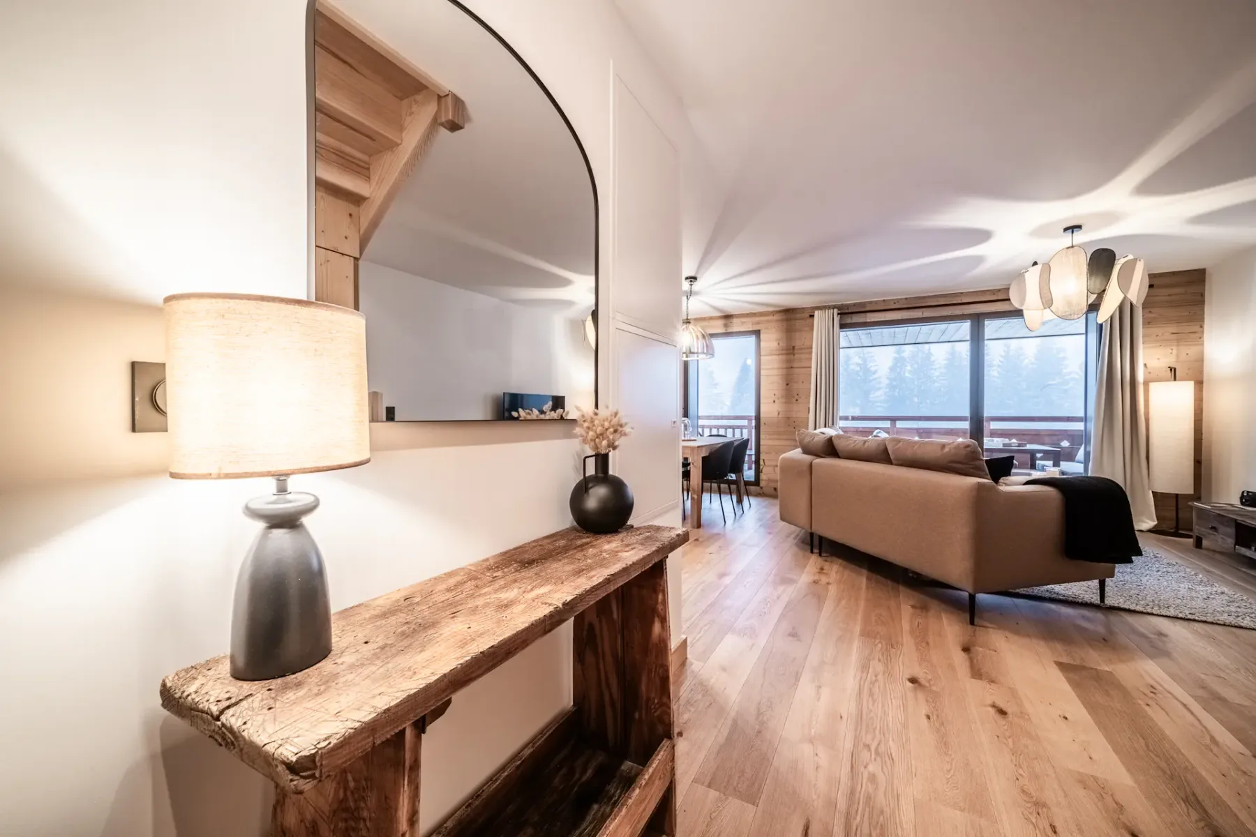 Sala de estar moderna con suelos de madera, un espejo grande y asientos acogedores. Hay una lámpara sobre una repisa rústica cerca de la entrada. Las grandes ventanas aportan luz natural y vistas a los árboles.