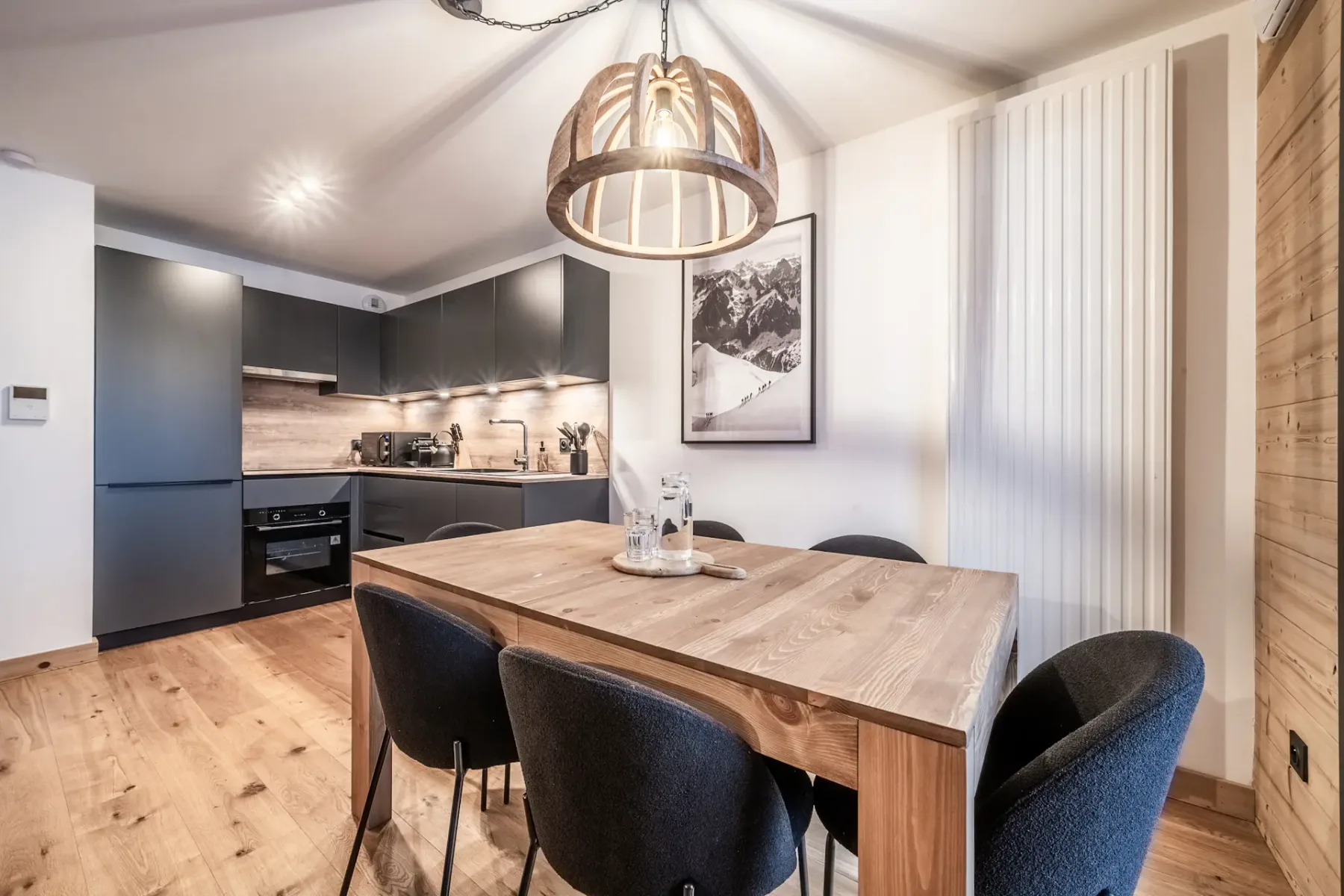 Cocina y comedor modernos con una mesa de madera, seis sillas negras, armarios oscuros y una gran lámpara colgante. Una fotografía de una montaña cuelga de la pared.