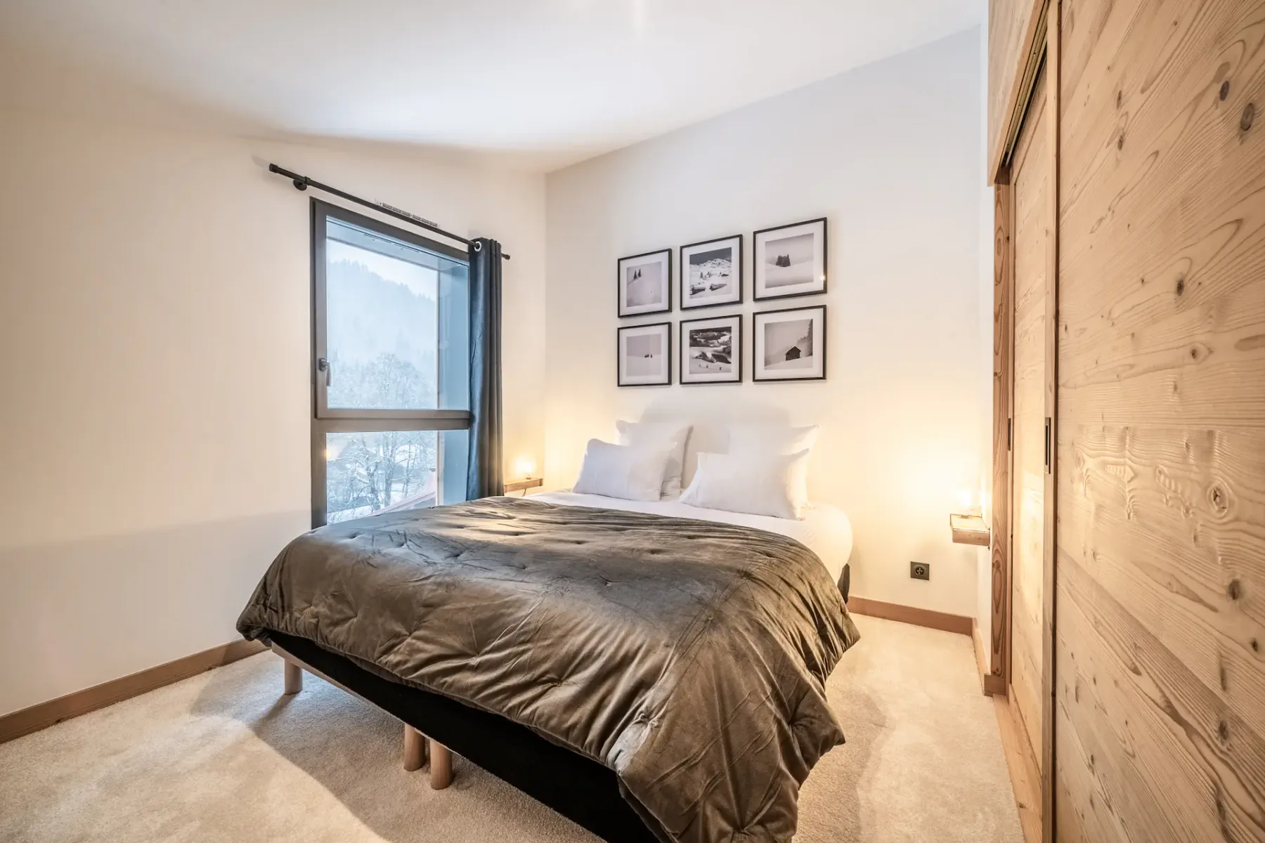 Dormitorio acogedor con una cama grande, un edredón gris y almohadas blancas. Hay una ventana con cortinas, cuadros enmarcados en la pared y una puerta corredera de madera. Una iluminación suave ilumina la habitación.