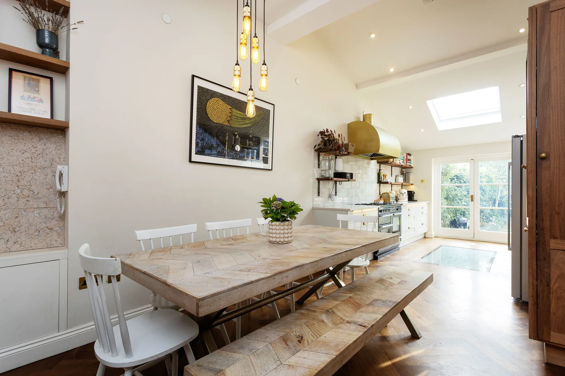 A bright dining area with a long wooden table and an open kitchen beyond.