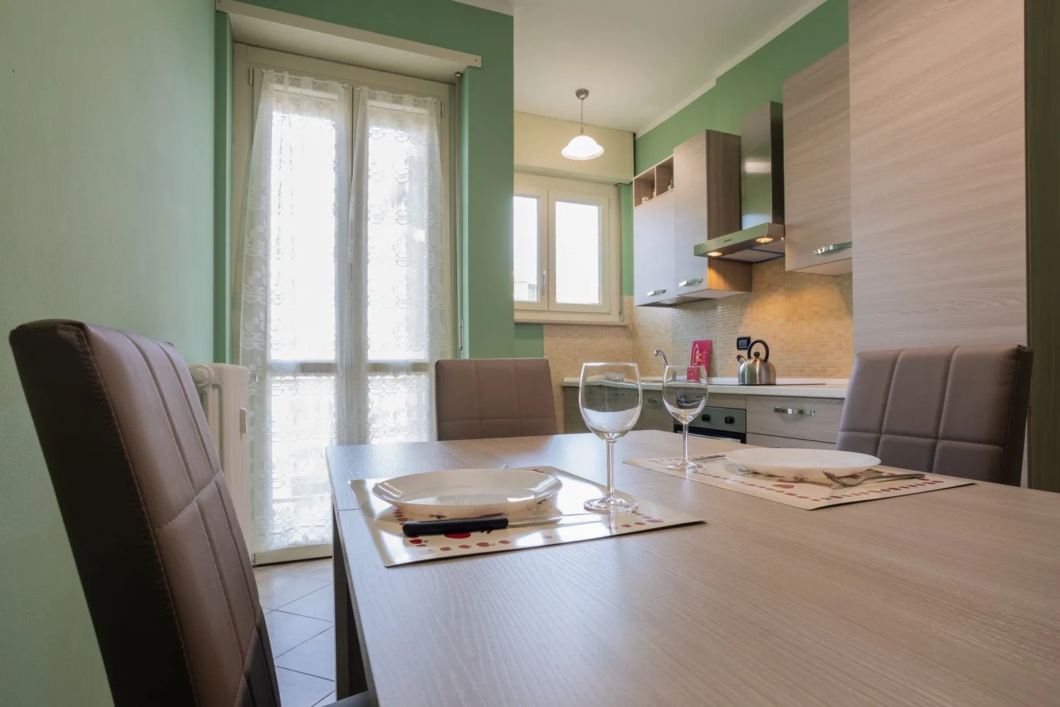 A dining table with place settings sits beside a compact kitchen with green walls.
