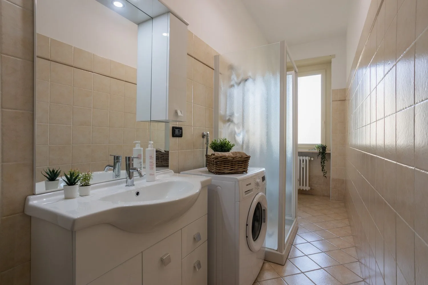 A tiled bathroom with a sink, washing machine, and glass shower.