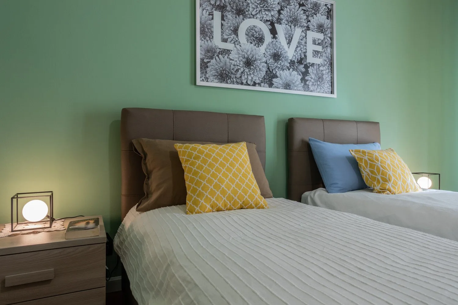 Un dormitorio con dos camas, cojines amarillos y una lámina floral enmarcada.