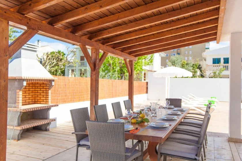 A covered outdoor dining area has a long table and a built-in brick barbecue.