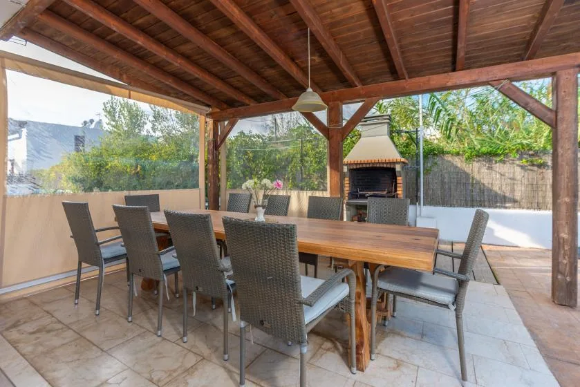 A covered outdoor dining area with a long table and chairs beside a barbecue.