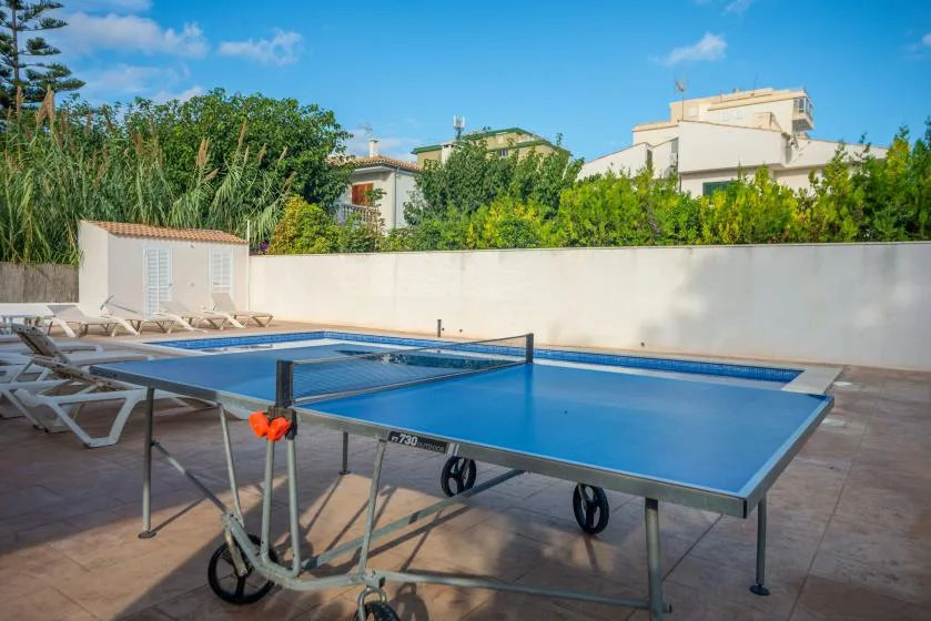 An outdoor table tennis table sits beside a swimming pool and sun loungers.