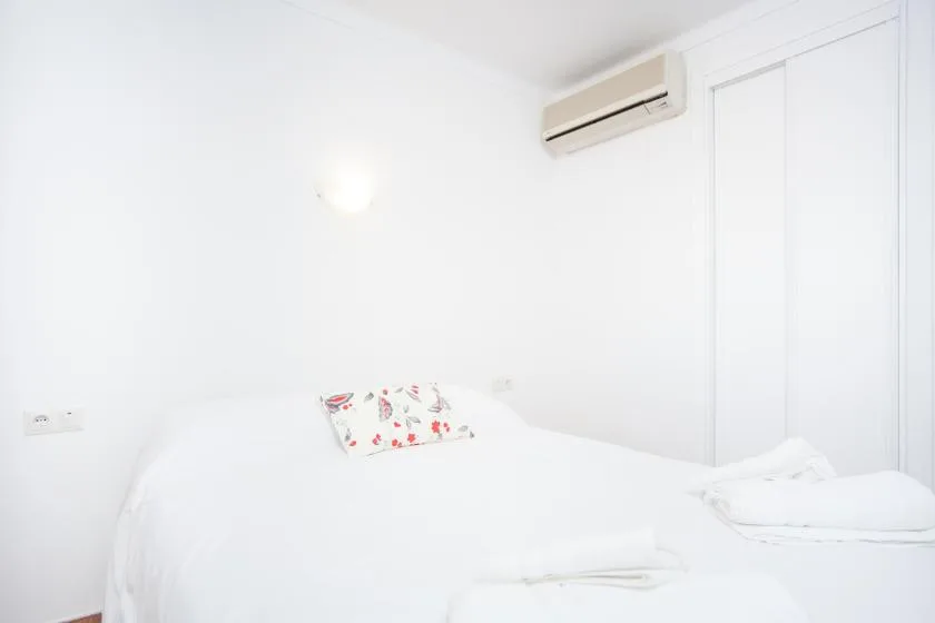 A bright bedroom with white bedding, a floral pillow, and a wall-mounted air conditioner.