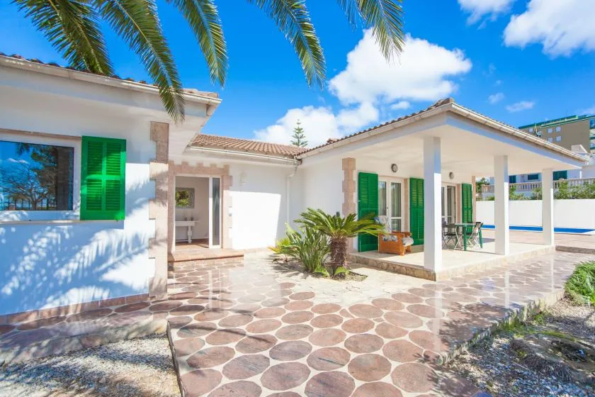 A white house opens onto a paved patio with green shutters and a covered seating area.